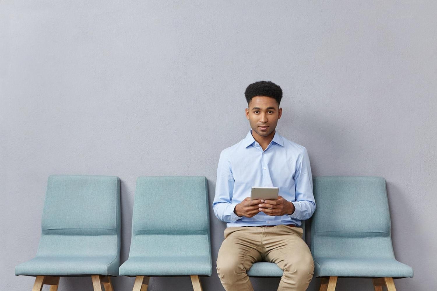 Young man seated in a modern waiting area using a tablet to track his queue ticket